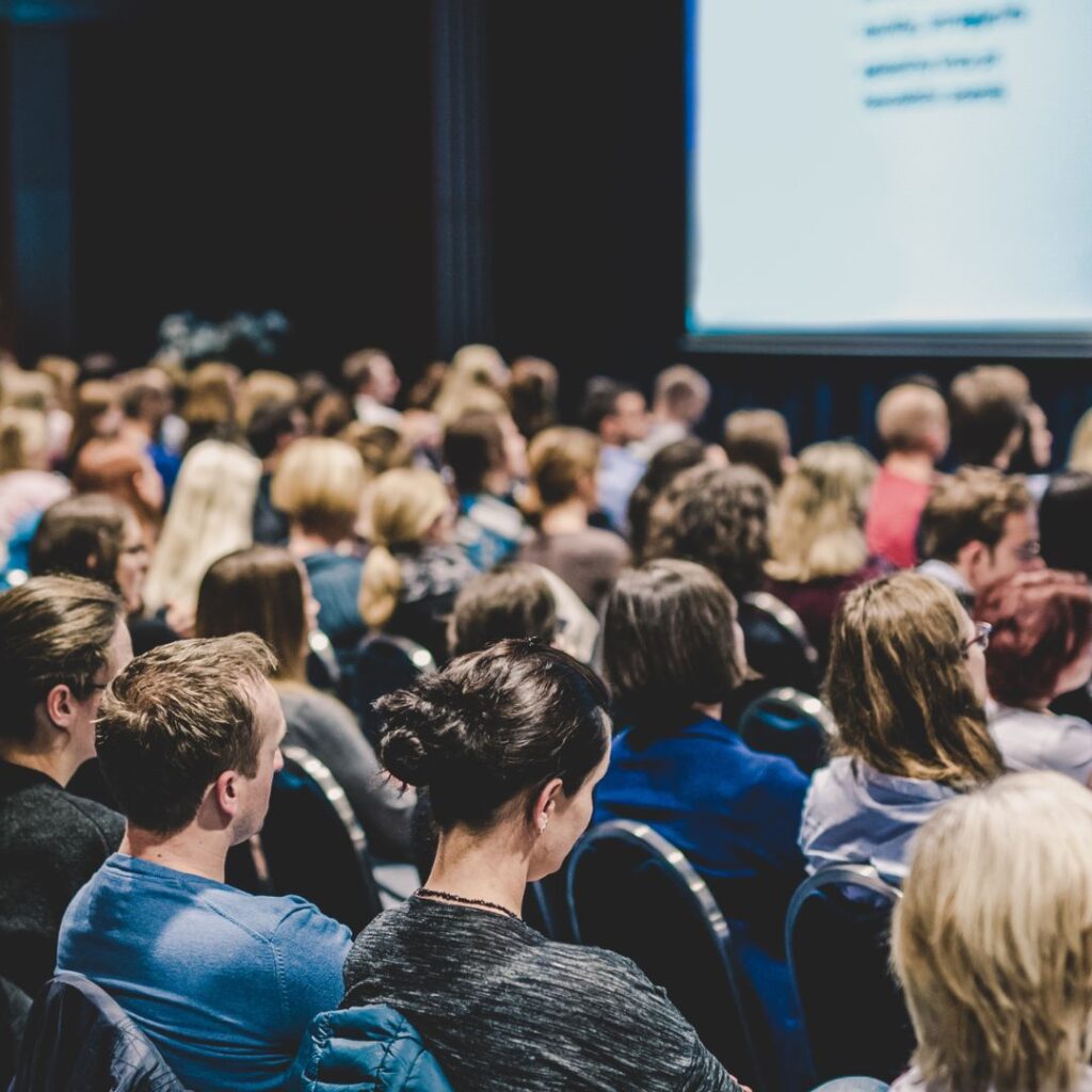 Vista desde atrás de una audiencia diversa en un auditorio mirando hacia una pantalla de presentación. Se ve una multitud de personas sentadas, con diferentes tonos de pelo y vestimenta en colores azules y negros.