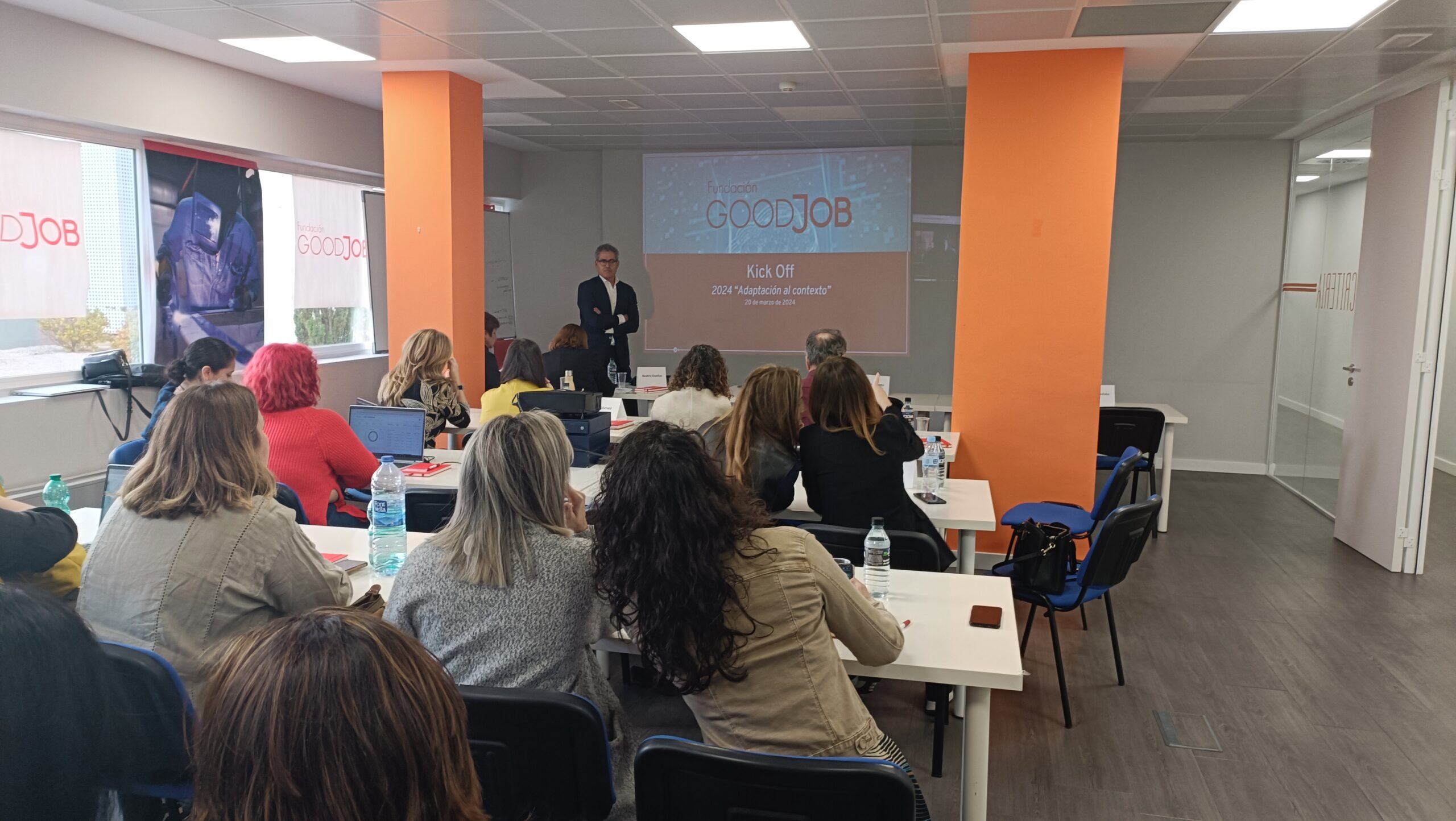 Sesión de formación de GoodJob donde varias personas están sentadas en mesas mirando hacia una pantalla que muestra la presentación 'GoodJob Kick Off'. Al frente hay un presentador de pie.