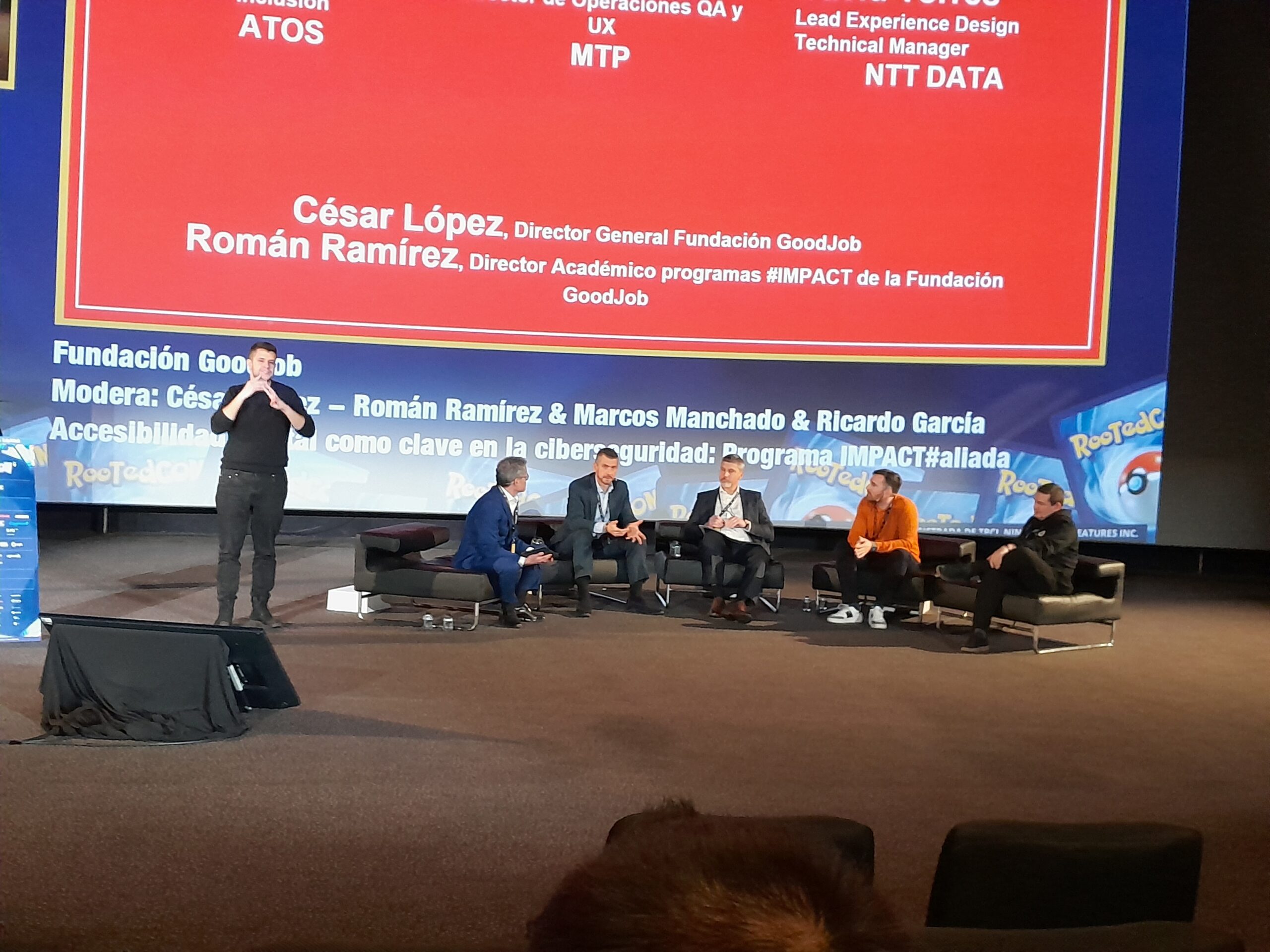 Mesa redonda en el evento RootedCON Madrid 2024 sobre accesibilidad y ciberseguridad. En el escenario hay cinco ponentes sentados y uno de pie. Se muestra un cartel con los logos de ATOS, MTP y NTT DATA junto con los nombres de César López y Román Ramírez de Fundación GoodJob.
