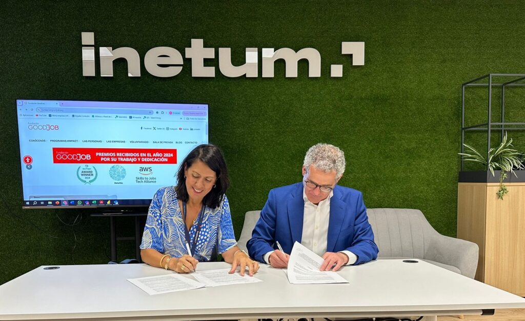 Dos personas firmando un acuerdo en una mesa, con el logotipo de Inetum en el fondo sobre una pared verde. En la pantalla detrás se muestra información de la Fundación GoodJob.