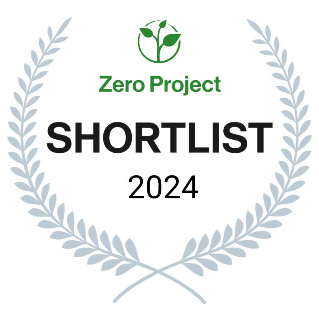 Emblema del Zero Project Shortlist 2024 que consiste en una corona de laurel gris con el logotipo verde del Zero Project en el centro, seguido del texto 'SHORTLIST 2024' en negro