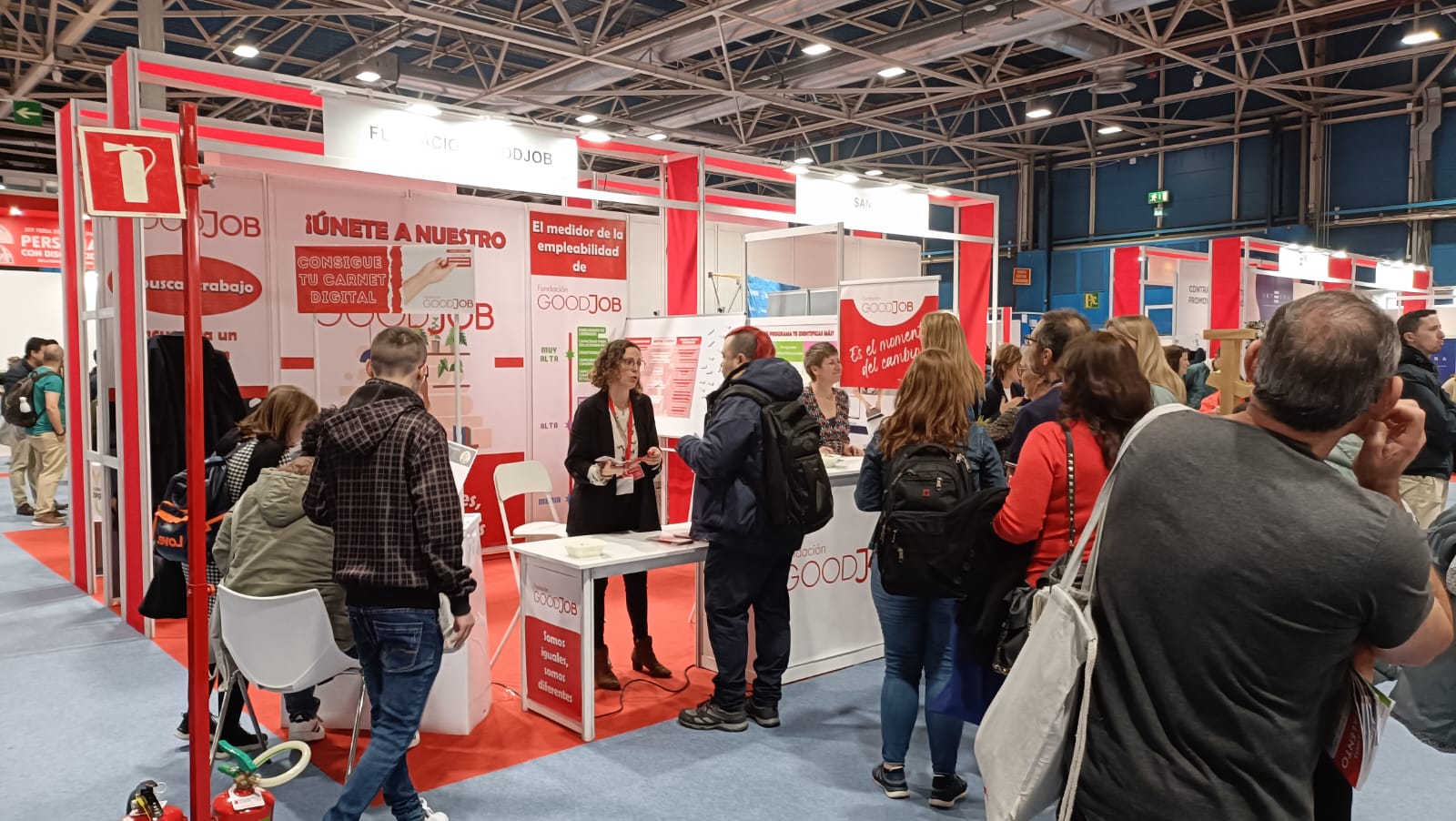 Stand de Fundación Goodjob en una feria o evento, con diseño en rojo y blanco, mostrando varios visitantes interactuando con el personal en los mostradores. El stand incluye carteles promocionales y el mensaje 'ÚNETE A NUESTRO' visible en una de las paredes