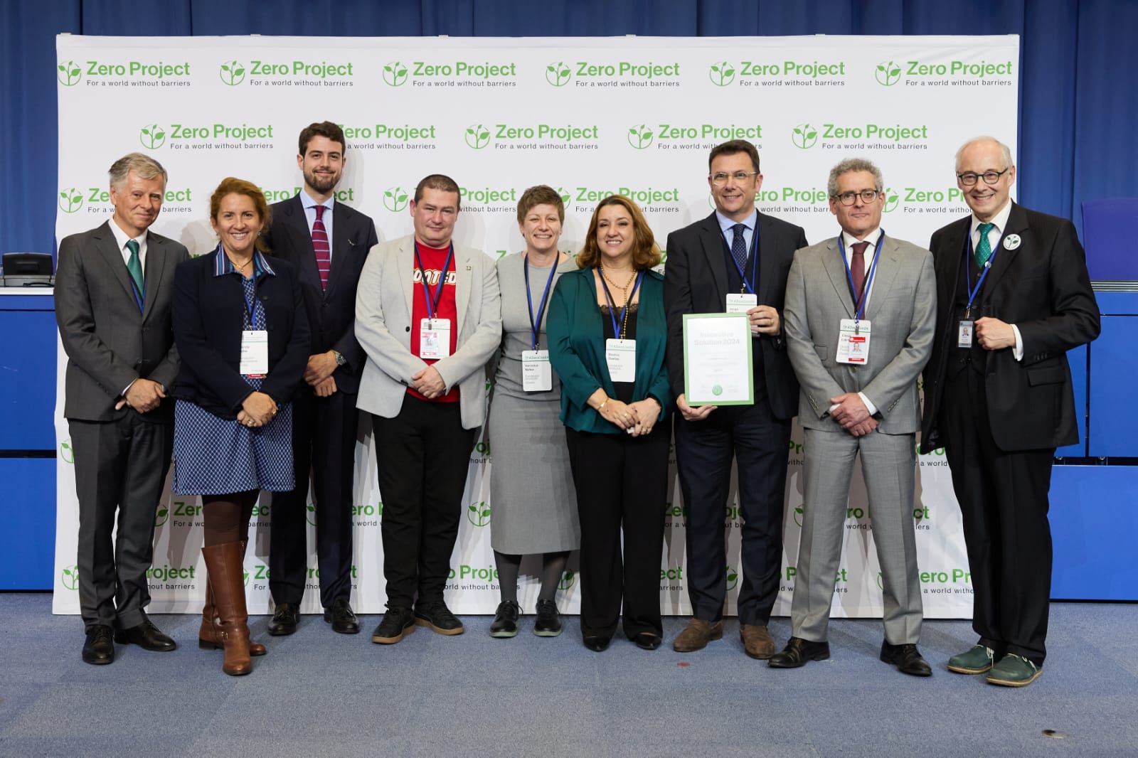 Grupo de nueve personas posando frente a un panel con el logotipo repetido de Zero Project. Una persona sostiene un documento y todos visten formal. El fondo incluye una pared azul y el suelo es gris