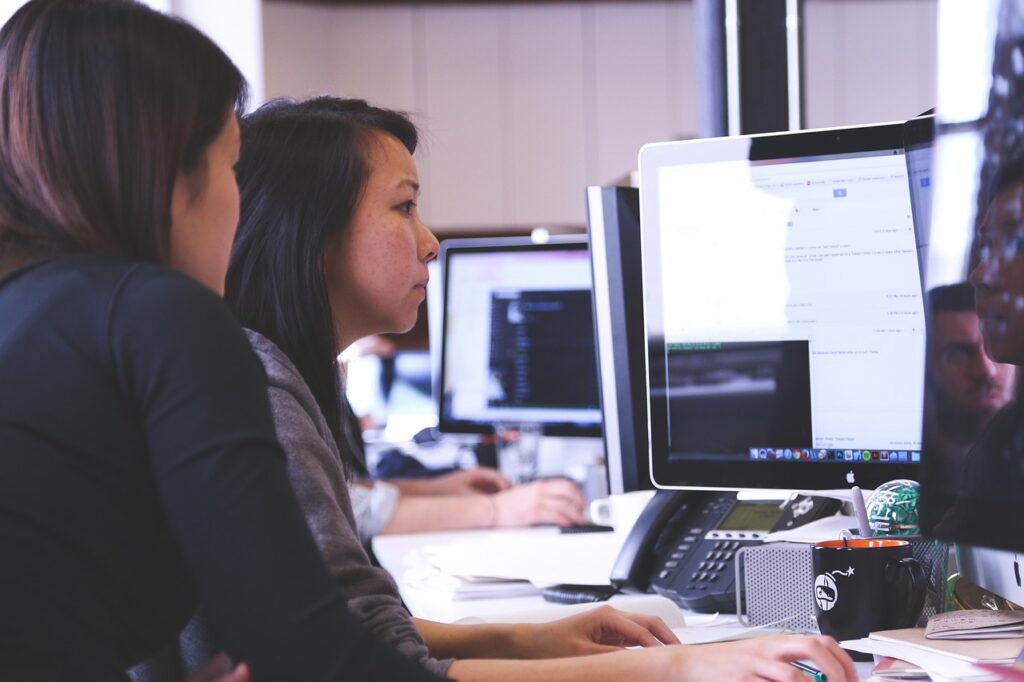 Dos mujeres en una oficina trabajando juntas frente a una computadora en un ambiente corporativo. Una está mirando la pantalla, mientras la otra está concentrada en el teclado