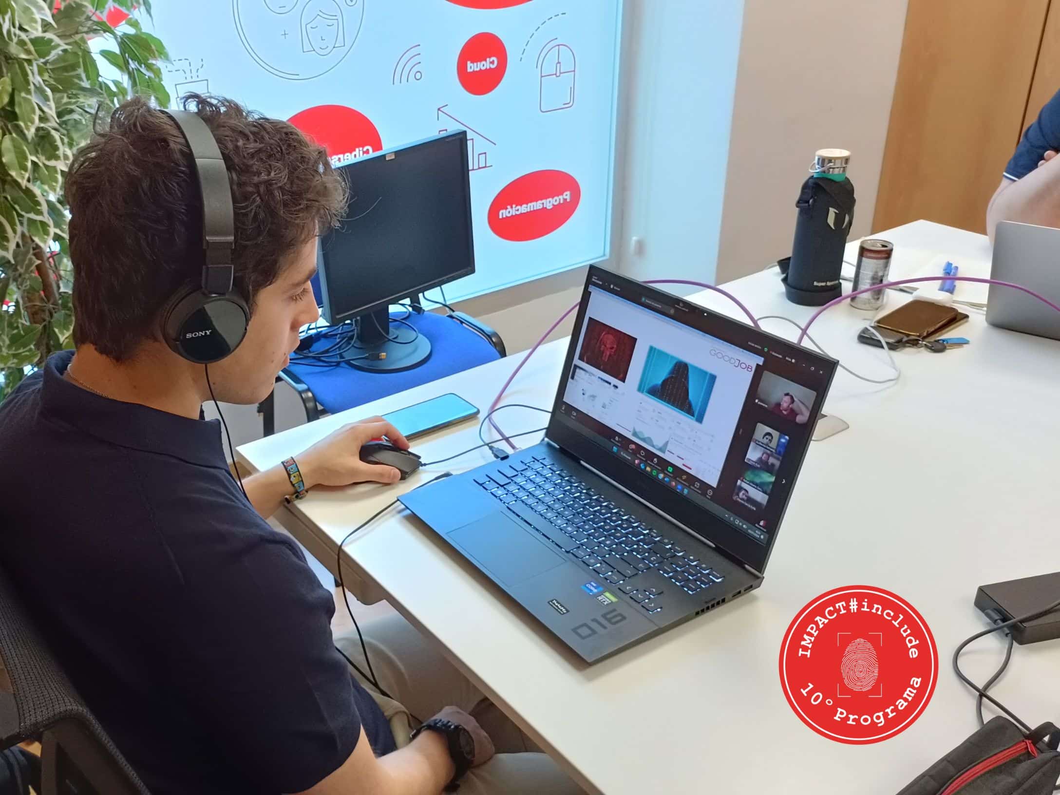 Joven con auriculares trabajando en un portátil durante una videollamada, con el logotipo del programa IMPACT#include visible en la esquina inferior derecha, en un espacio de coworking decorado con elementos tecnológicos.